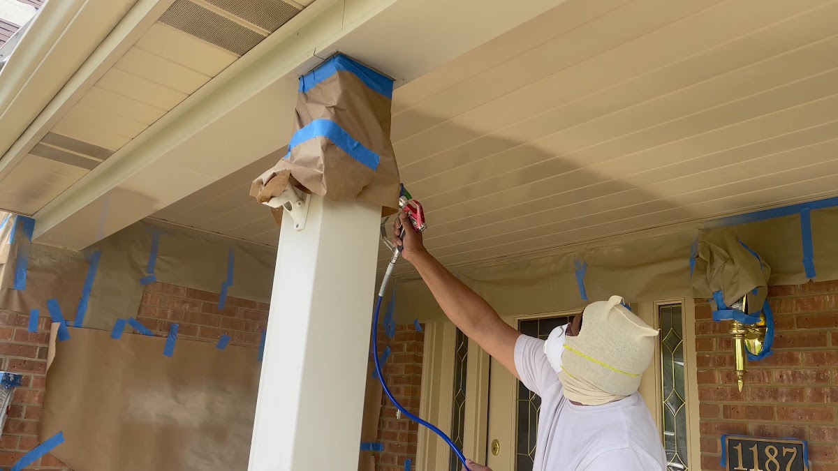 Exterior spray-painting of a porch column and soffit, with blue painter's tape masking visible